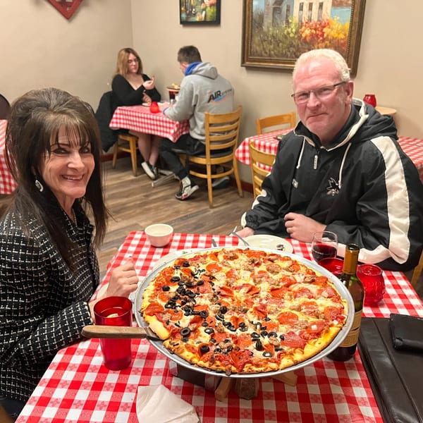 A couple smiles at their table with a large pizza, red wine, and checkered tablecloths at Italian To Go.