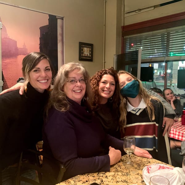 Four women smiling at the camera, with a large Italian-themed mural behind them.