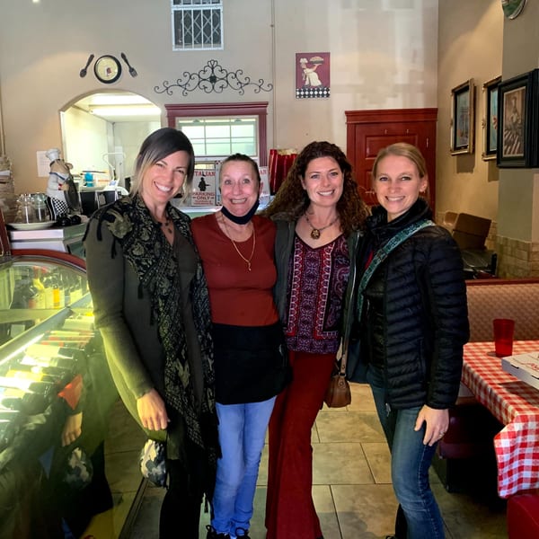 Four smiling women pose inside Italian To Go / Bit Of Italy restaurant, with checkered tablecloths and decor.