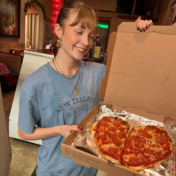 Smiling young woman in a restaurant holding an open pizza box with two small pepperoni pizzas.