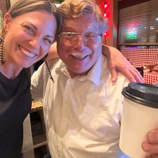 A smiling woman and man with glasses pose together, holding a coffee cup in an Italian restaurant.