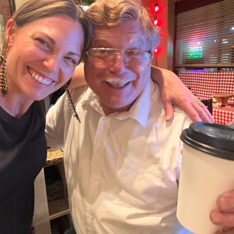 A smiling woman and man with glasses pose together, holding a coffee cup in an Italian restaurant.