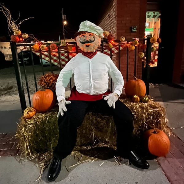 A chef scarecrow with a pumpkin head sits on hay bales, surrounded by pumpkins and fall decor.