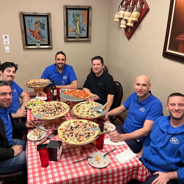 Seven men in blue shirts enjoying multiple pizzas and drinks at a red and white checkered table.