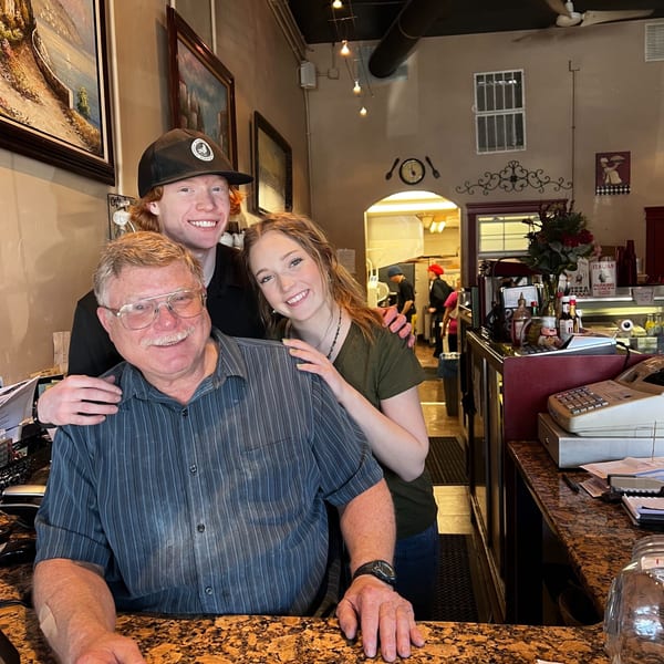 Three smiling people, two young and one older, stand together behind a counter at Italian To Go.