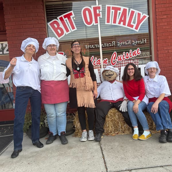 Five people and a scarecrow in chef hats and costumes pose outside "Bit Of Italy" restaurant.