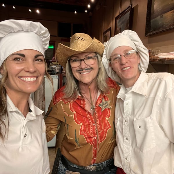 Three smiling women, two in chef hats and one in a cowboy hat and mustache, pose together.