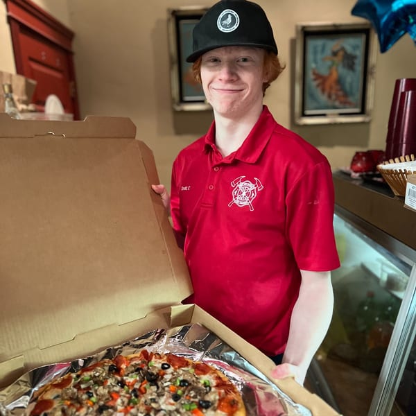 Smiling young man in a red shirt and black hat holding an open box with a large pizza.