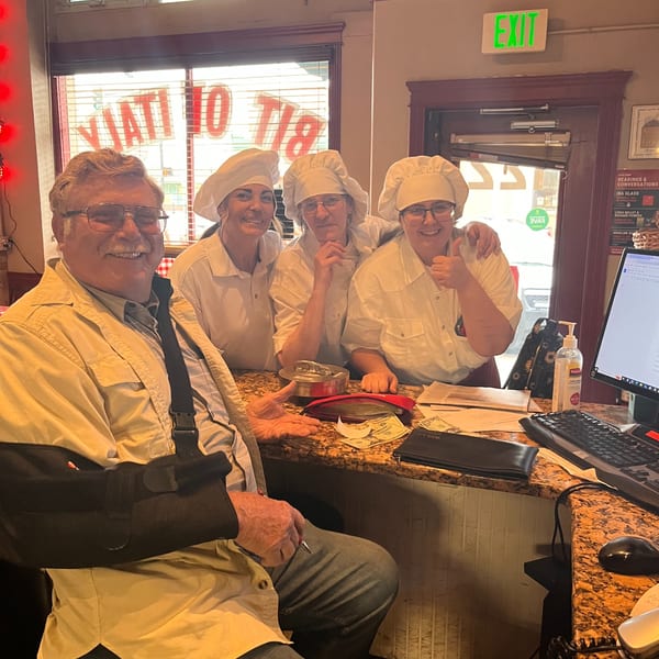 A man with a sling sits at a counter with three smiling Italian To Go staff in chef hats.