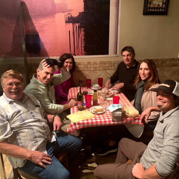 Six happy diners enjoying Italian food and wine at a table with a red checkered tablecloth.