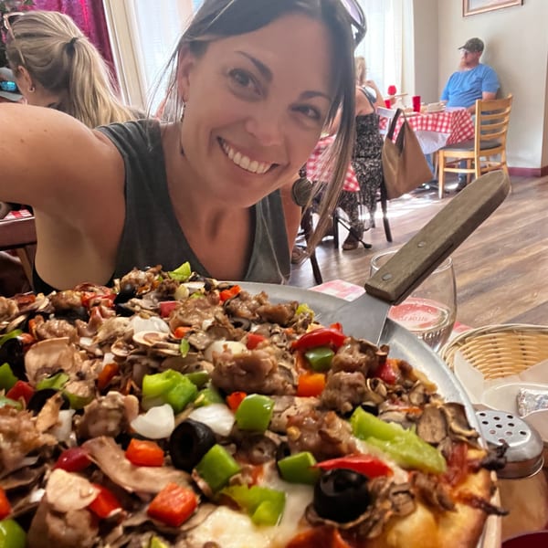 A smiling woman takes a selfie with a large, loaded Italian pizza in a restaurant setting.