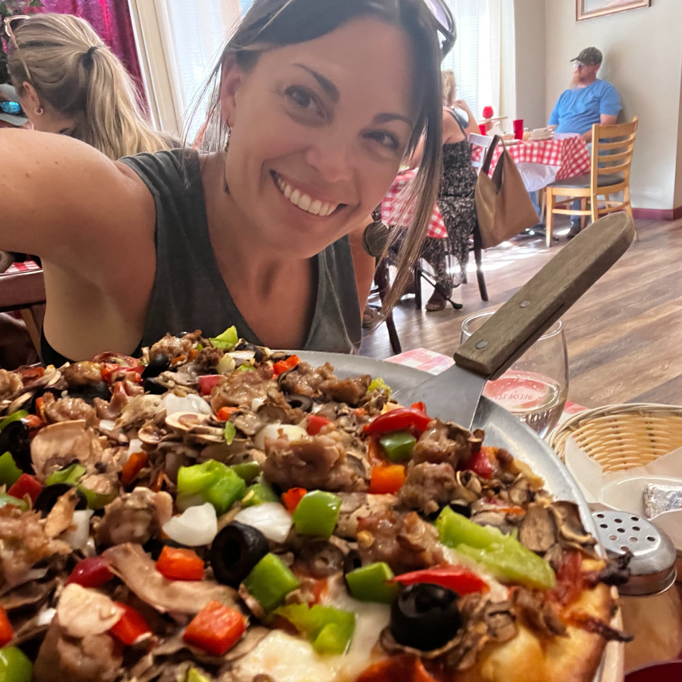A smiling woman takes a selfie with a large, loaded Italian pizza in a restaurant setting.