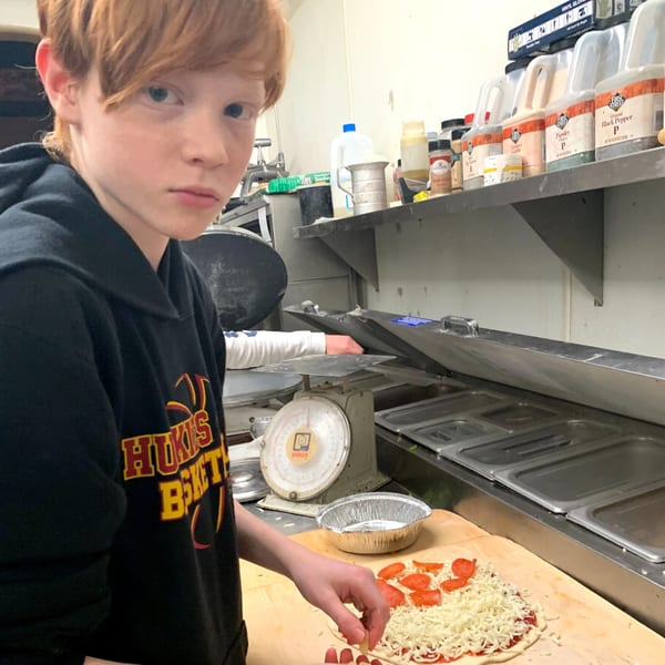 A young person with red hair making a pepperoni pizza in a commercial kitchen.