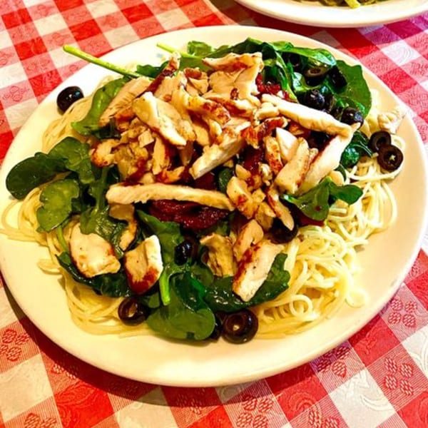 Italian pasta dish with chicken, spinach, sun-dried tomatoes, and black olives on a red and white checkered tablecloth.