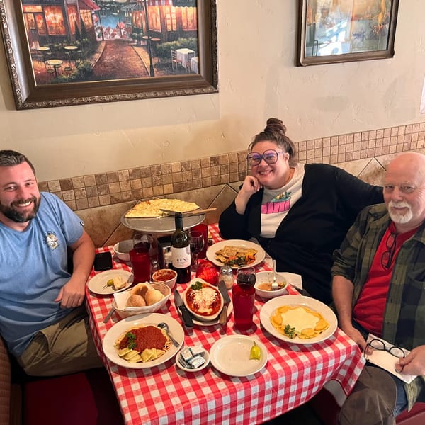 Three happy diners enjoying Italian food, including pizza and pasta, at a table with a red checkered tablecloth.