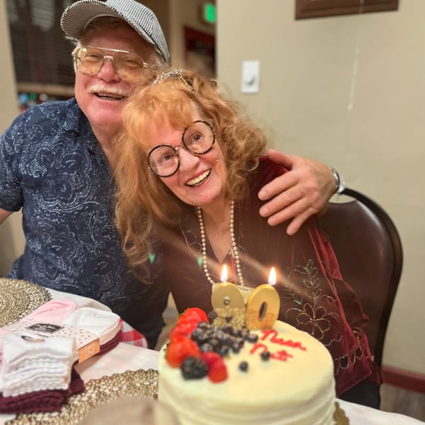 A smiling couple celebrates a birthday with a cake topped with berries and "90" candles.