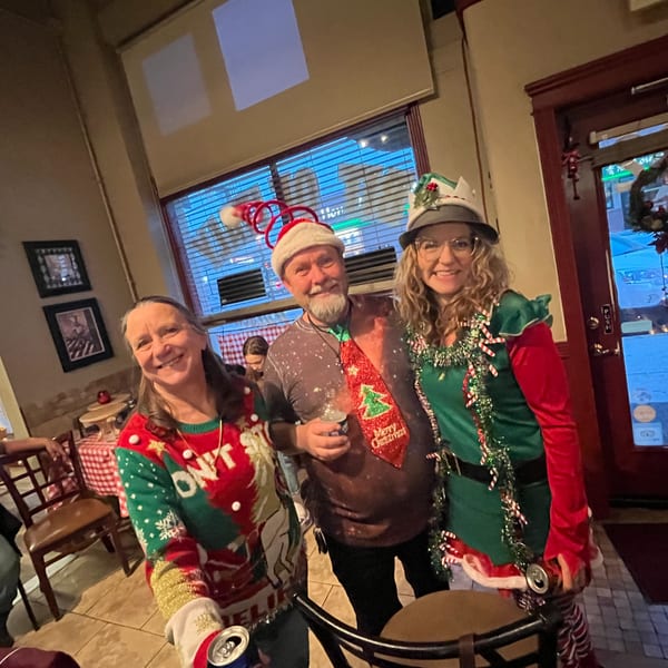Three smiling people in festive Christmas attire, including an elf costume and Santa hats, at Italian To Go.
