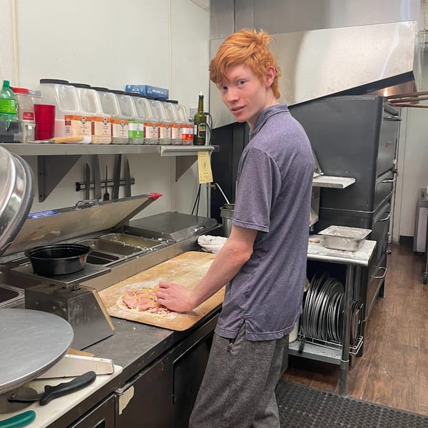 A red-haired chef smiles while preparing an Italian pizza with ham and cheese in a kitchen.