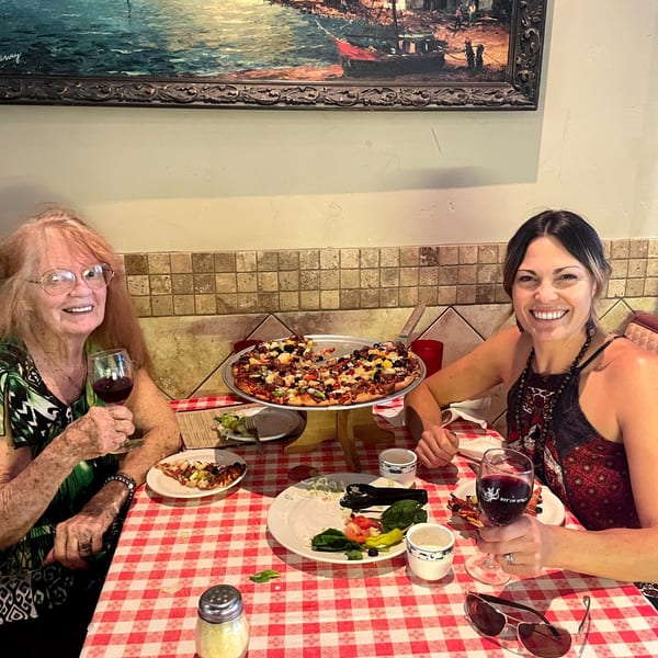 Two women smiling at a table with a large Italian pizza and red wine.