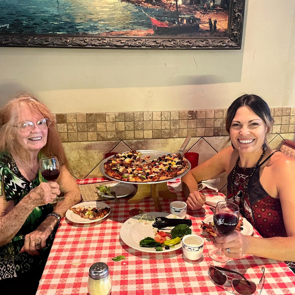 Two women smiling at a table with a large Italian pizza and red wine.
