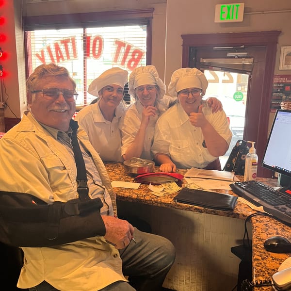 A man with a sling poses with three smiling women in chef hats behind a counter at Italian To Go.